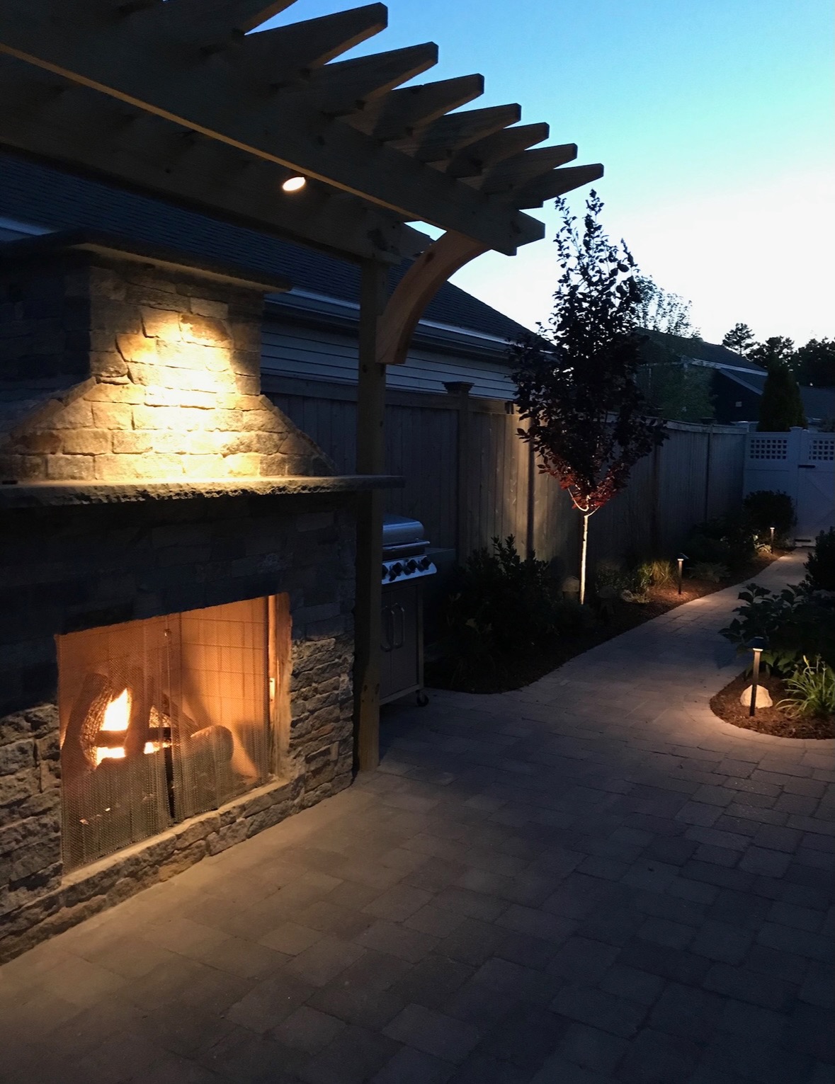 Outdoor stone fireplace with pergola lit at dusk alongside a paver patio